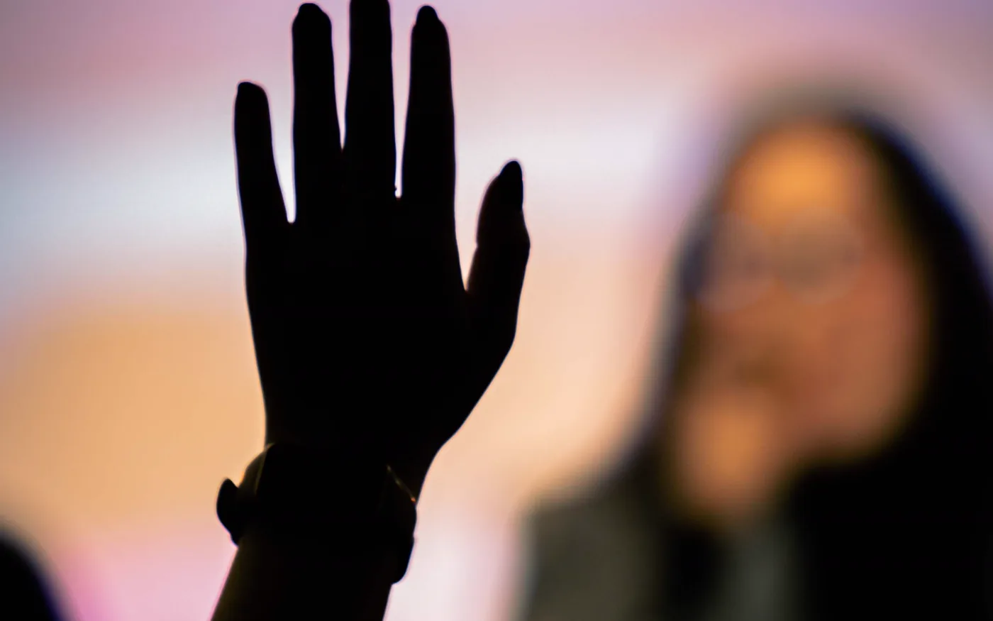 A raised hand silhouetted against a lit screen, waiting to ask a question