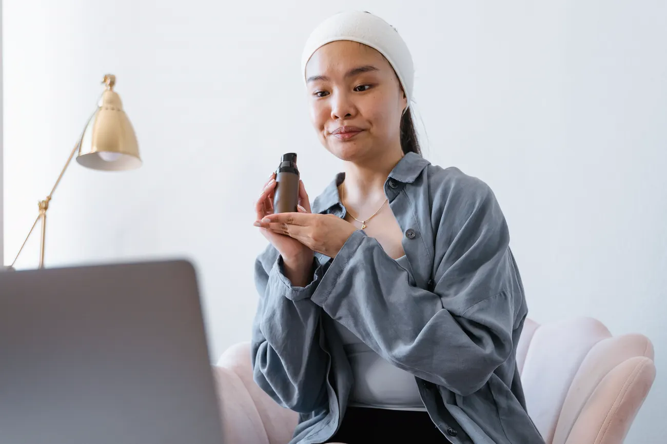 A woman holding a skincare bottle up to a laptop camera during a video call