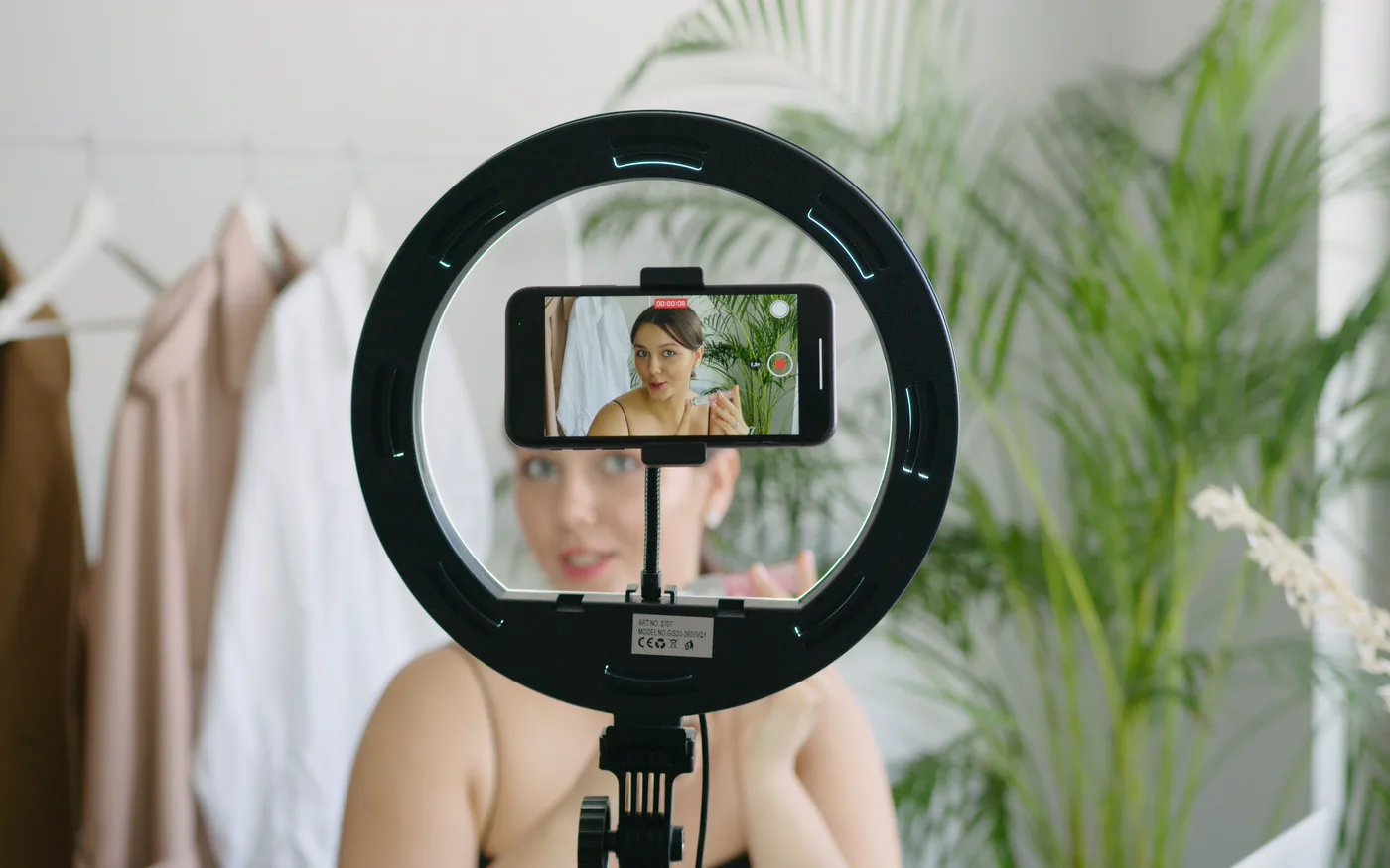 A smartphone in a ring light filming a woman beside a rail of clothes