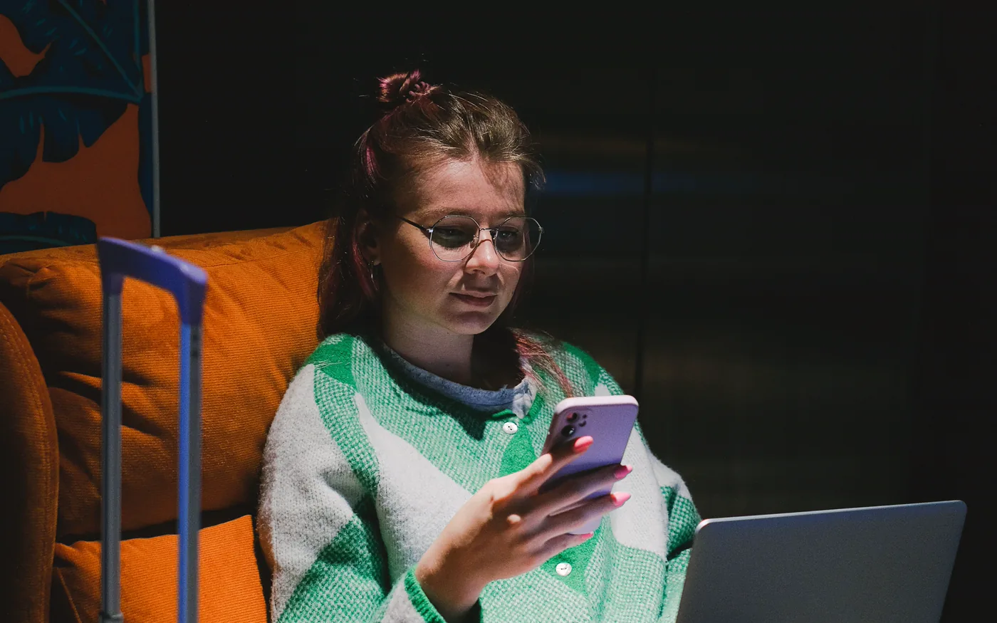 A woman in an armchair at night, checking her phone with a laptop open on her knees